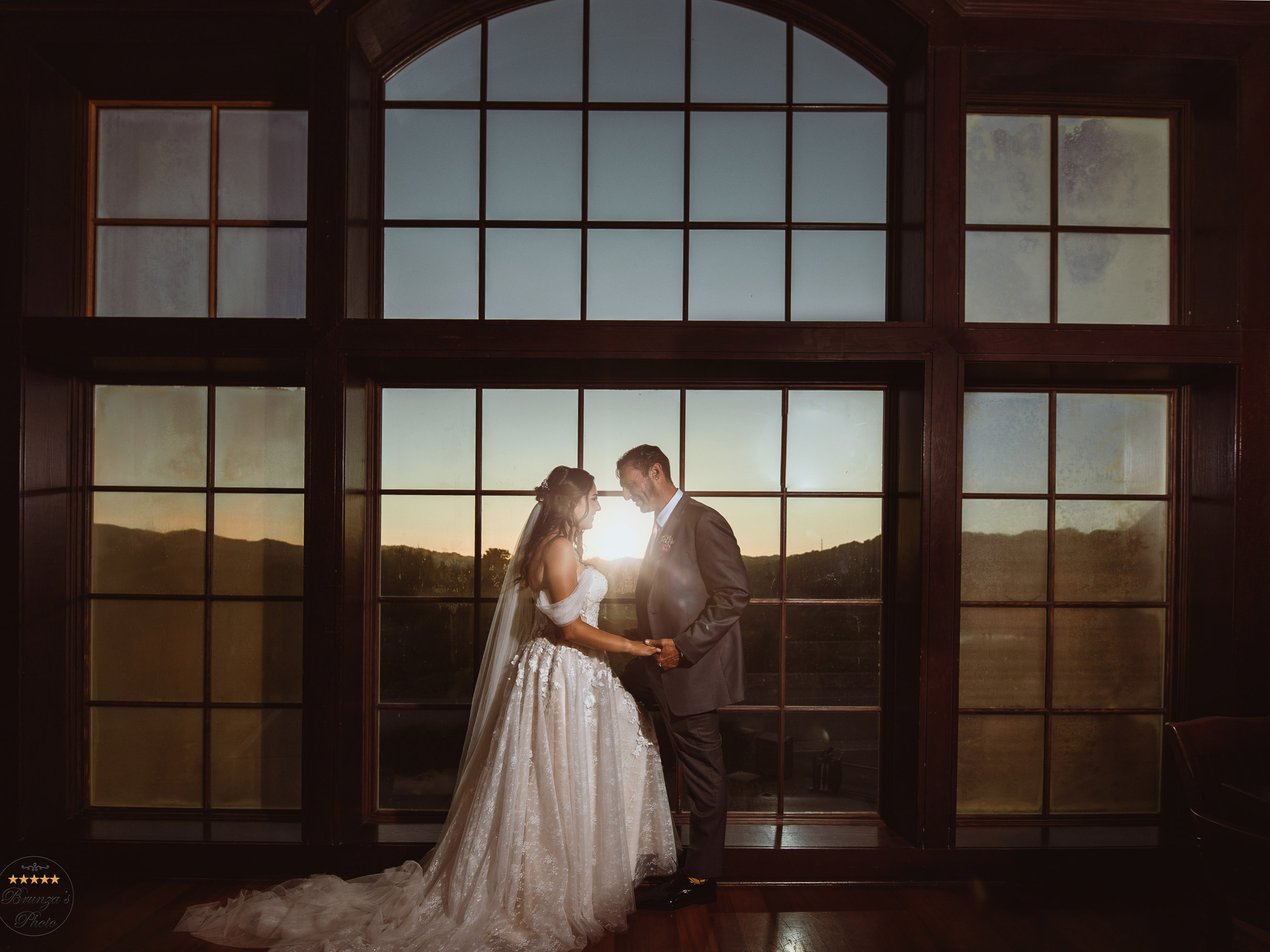 A bride and groom standing in front of a window at sunset.