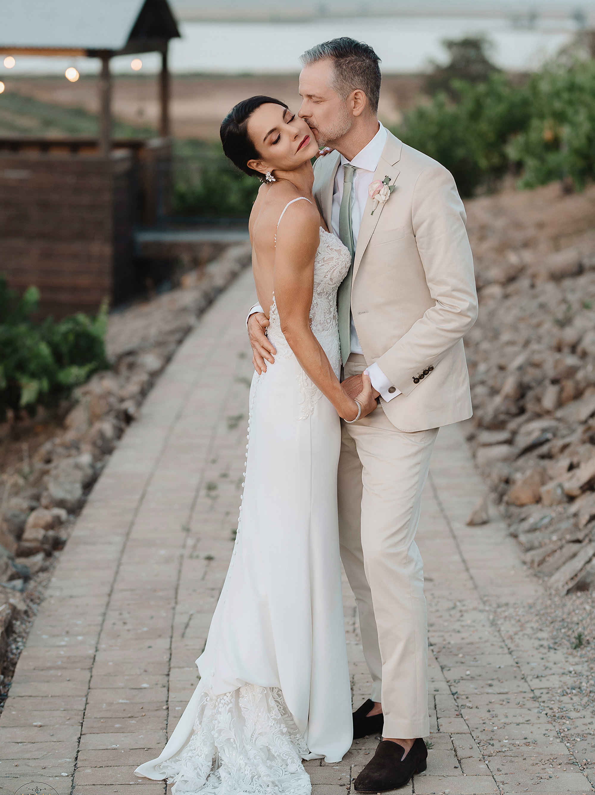 A bride and groom kissing on a pathway in front of a vineyard.
