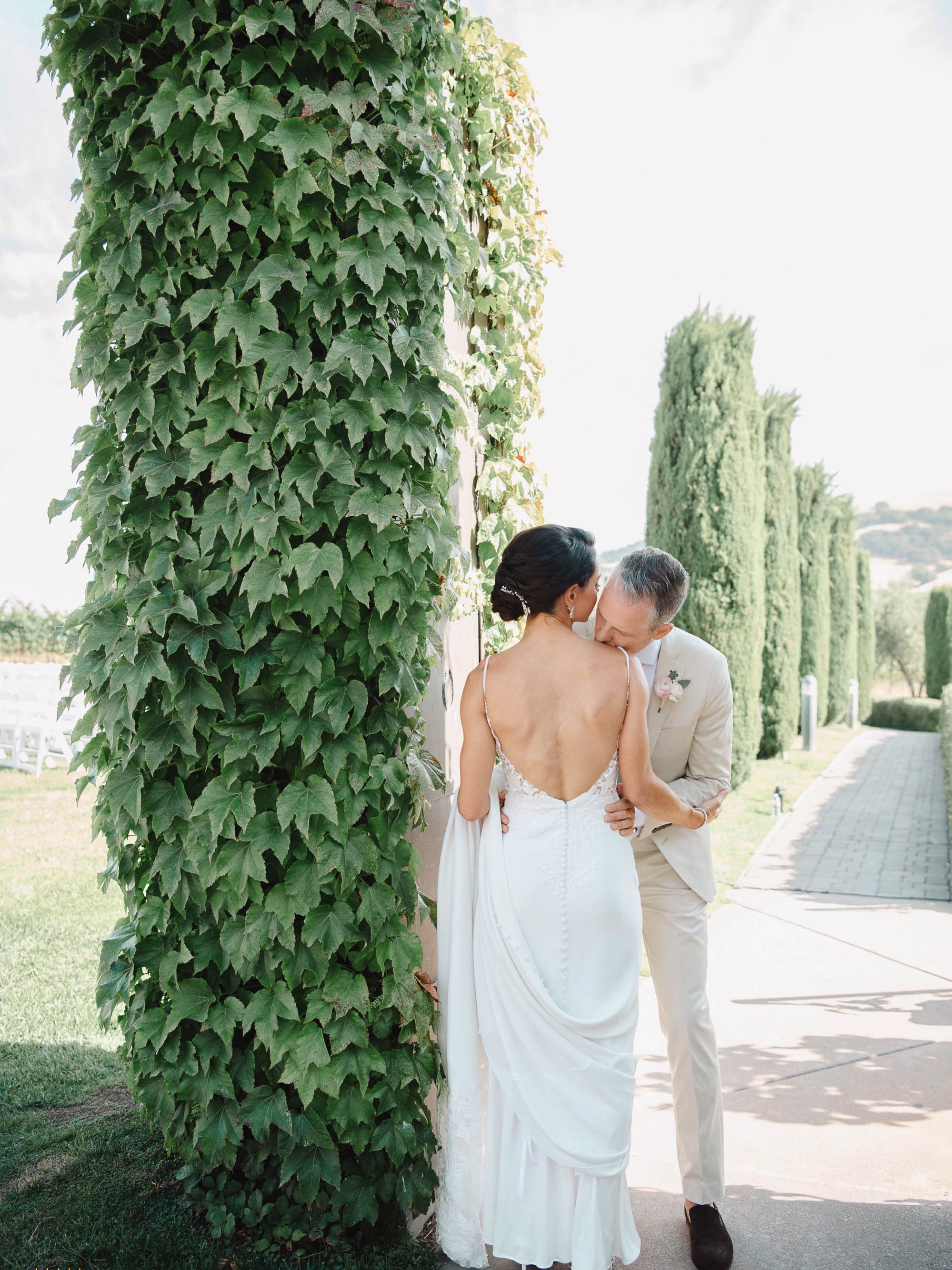 A bride and groom kiss in front of an ivy covered arbor.